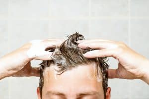 young man washing his hair, taking a shower with foam on his head holds fingers in hair in bathroom